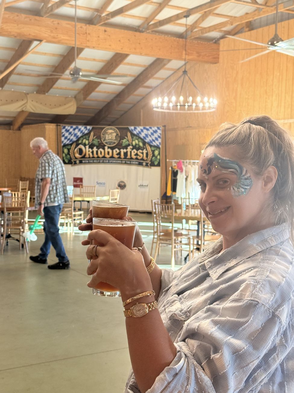A woman with blue face paint around one eye toasting with a pint in the Big Barn, the Oktoberfest backdrop behind her