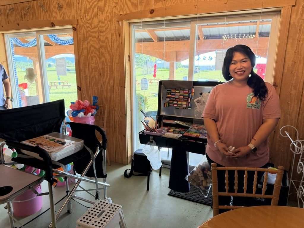 The face painter smiling beside her open paint kit inside the barn, balloon flowers behind her
