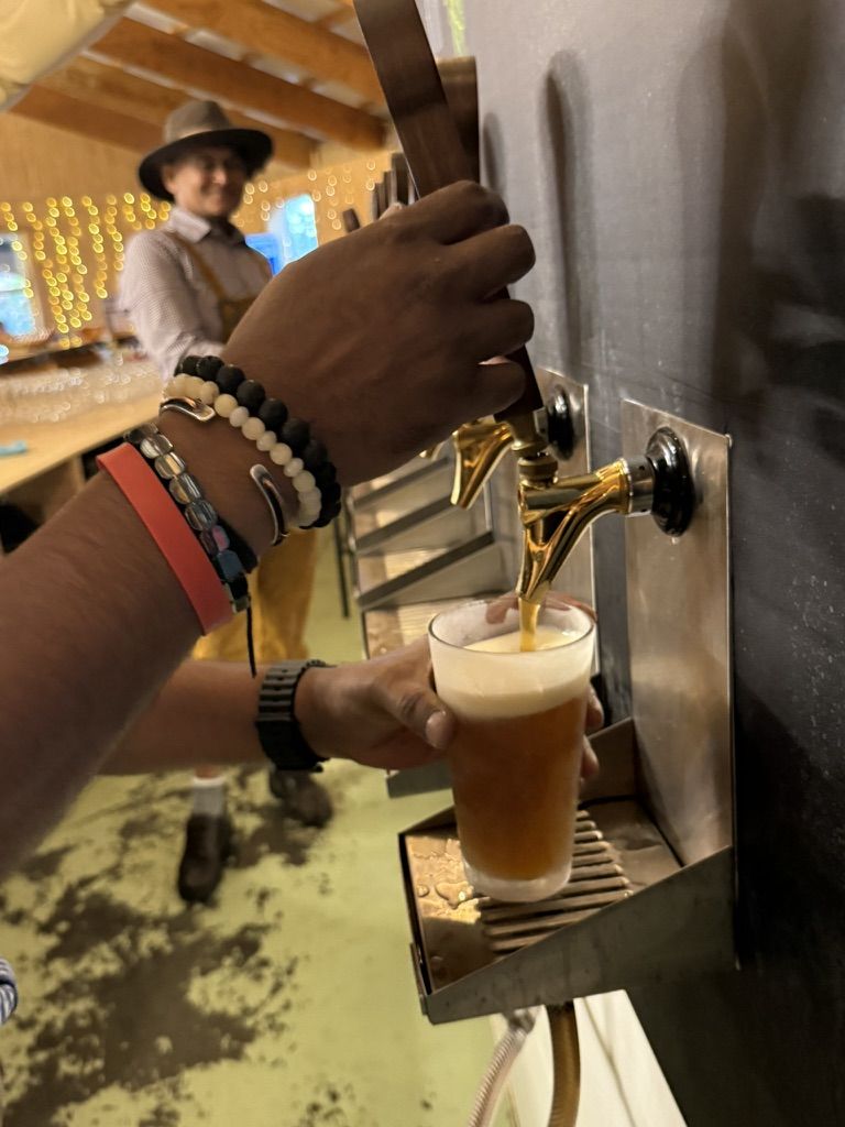 A hand pulling a brass tap to fill a pint glass, a man in lederhosen smiling in the background