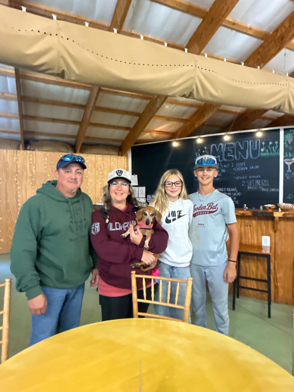 Four guests standing by the chalkboard menu, one holding a small dachshund in a pink sweater