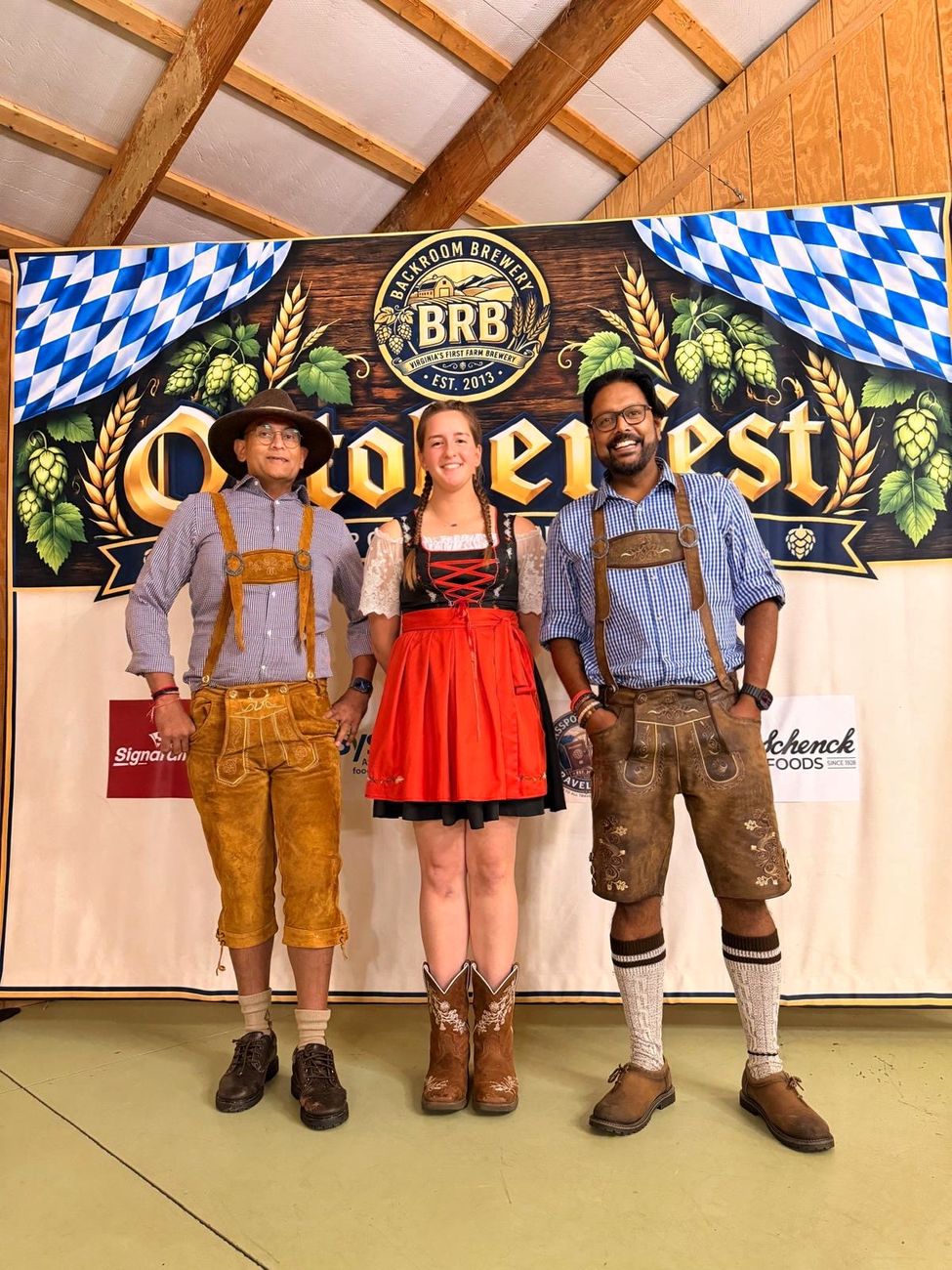 Two men in lederhosen and a woman in a dirndl and cowboy boots standing in front of the Oktoberfest backdrop