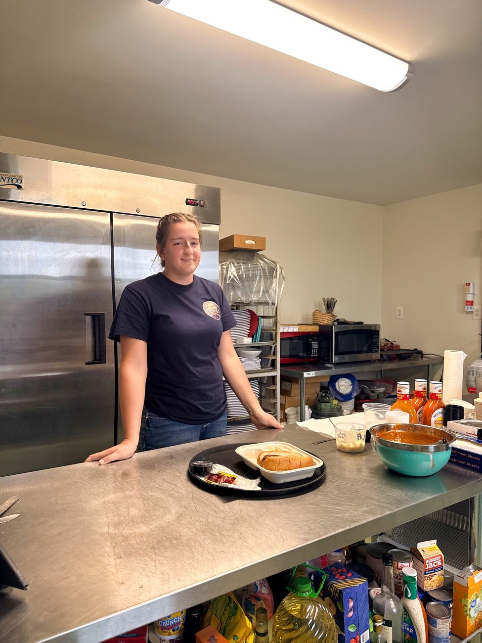 A team member in the kitchen behind a tray holding a brat on a bun