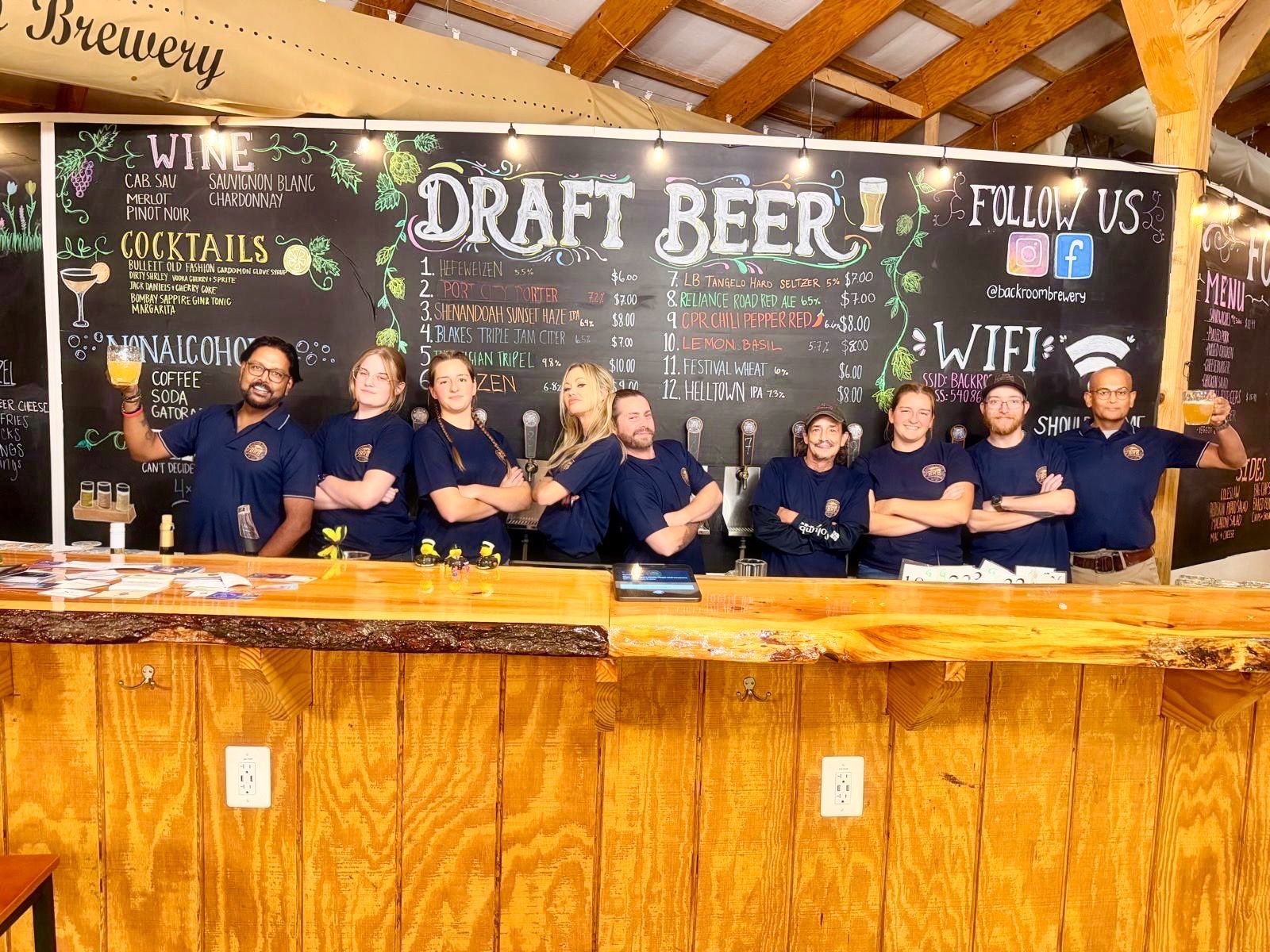 Nine Backroom Brewery team members in navy shirts lined up behind the bar in front of the chalkboard draft list, two raising mugs