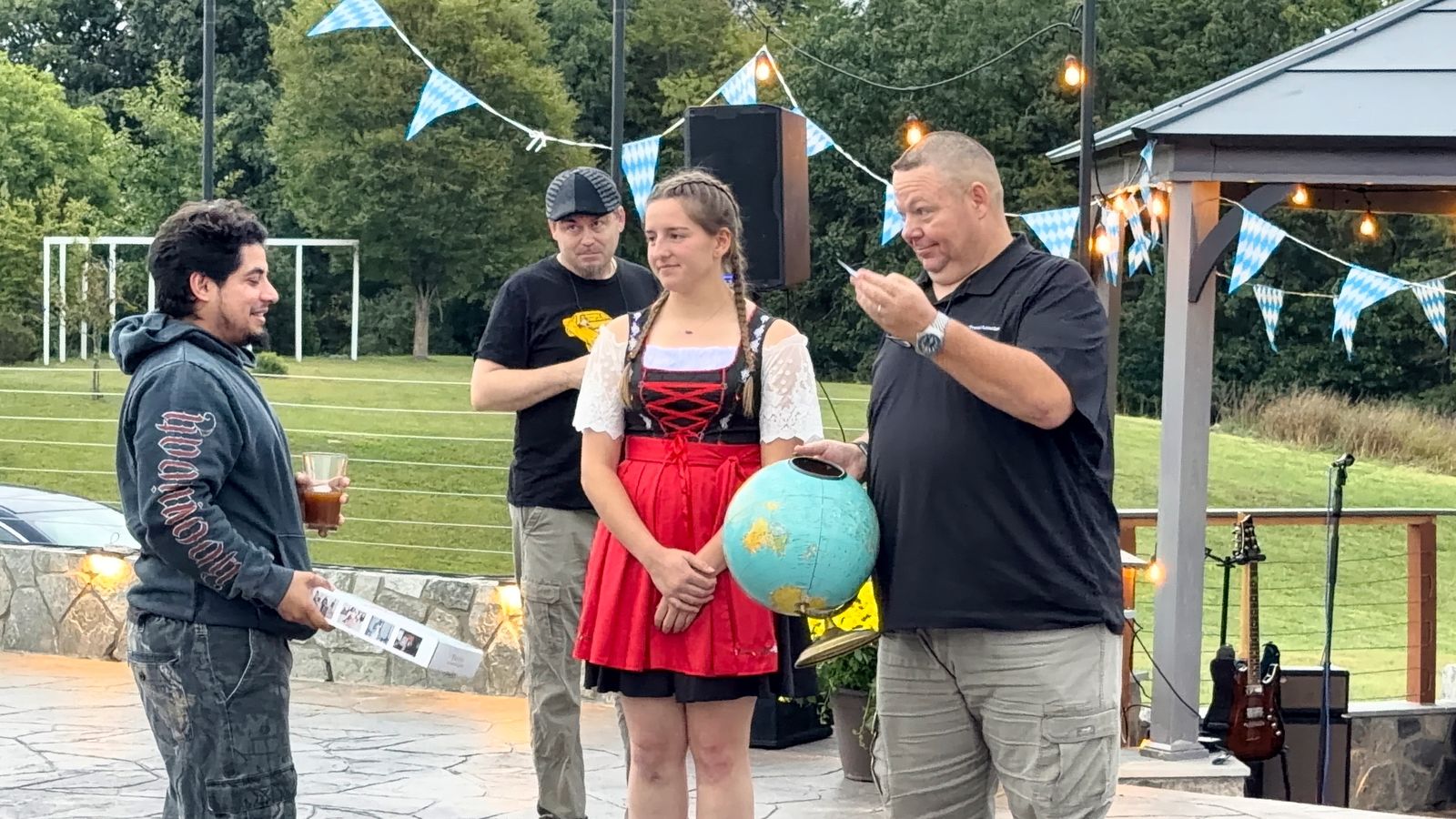 A man drawing a slip of paper from a globe on the patio while a woman in a dirndl and two guests look on