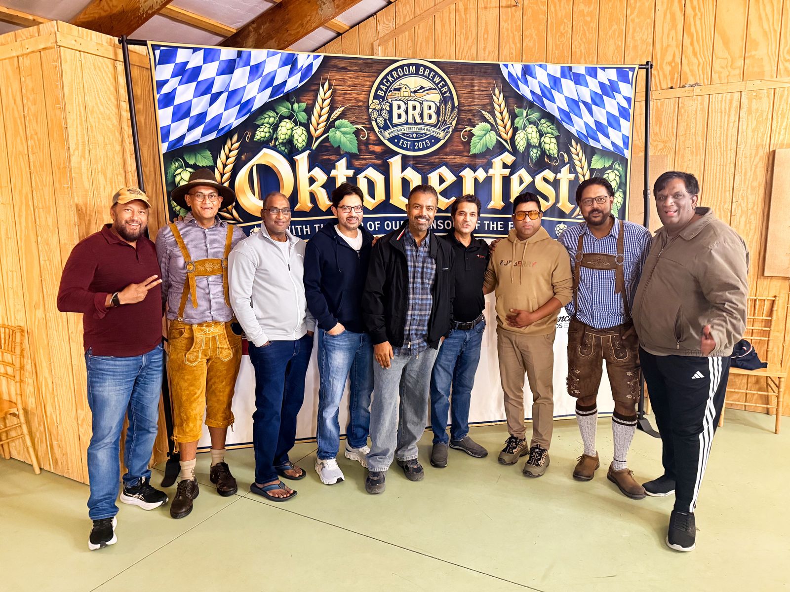 Nine men, two in lederhosen, lined up in front of the Backroom Brewery Oktoberfest backdrop