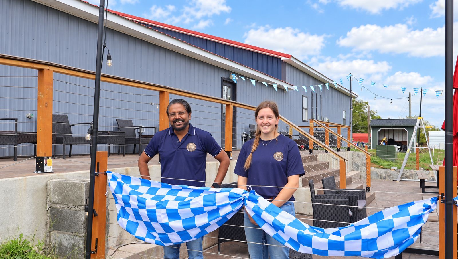 Two Backroom Brewery team members in navy BRB shirts standing behind blue-and-white bunting on the patio rail