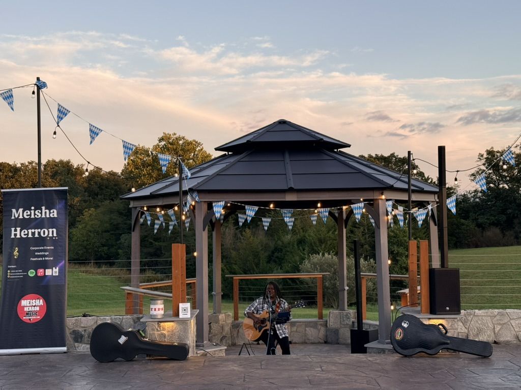 Meisha Herron playing acoustic guitar in front of the gazebo at sunset, bunting strung overhead