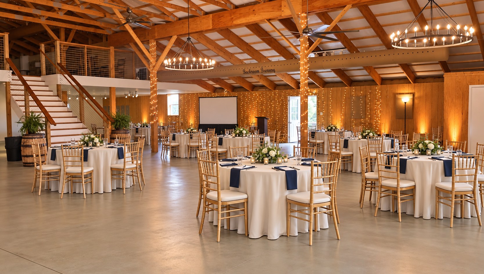 The Big Barn set with round tables, gold Chiavari chairs, a projection screen and a podium